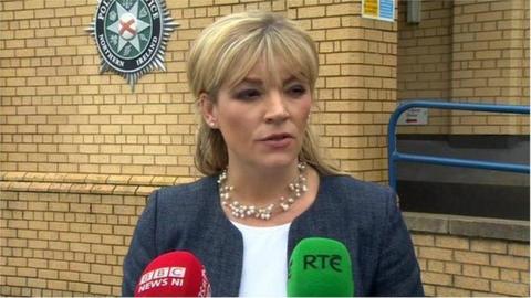 Una Kelly, who has blonde hair cut into a fringe and wears a grey blazer over a white top, talks to the media outside a PSNI police building. BBC and RTE branded microphones are visible in front of her