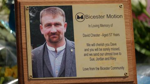 A gold plaque among the flower tributes. It includes an image of Dave Chester and a logo for Bicester Motion. It says: "We will cherish you Dave and you will be sorely missed, as we send our utmost love to Sue, Jordan and Riley. Love from the Bicester Community"