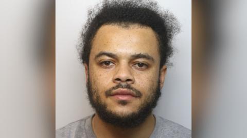 A mugshot of Oliver Onyeahasi. He has brown eyes, short dark curly hair and facial hair. He is pictured against a plain background in a grey t-shirt. He is looking directly at the camera with a blank expression.