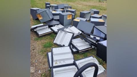 The fridges, dumped on the grassed area. Most of the doors have separated from the bodies of the fridges. They are a dark grey colour.