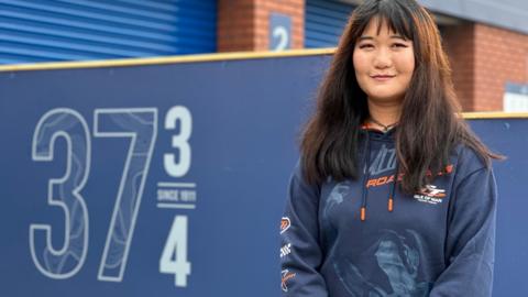Julie Sun, who has long dark hair and is wearing a blue TT branded hoodie. She is standing next to a large sign with 37 3/4 on it.