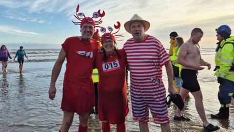 Three swimmers pose for the camera - a man and a women are wearing red crab outfits with one man wearing a red and white striped Victorian bathing costume with hat