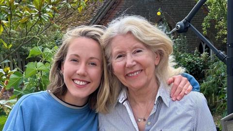 Frannie and Christine Brown together with arms round one another. There is green shrubbery in the background.