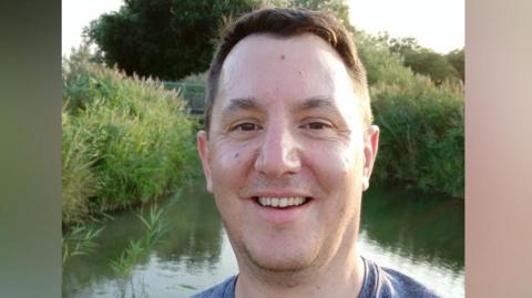 James Clements is standing in front of a small lake which is surrounded by tall grass. He has short brown hair, and is smiling at the camera. He is wearing a blue T-shirt.