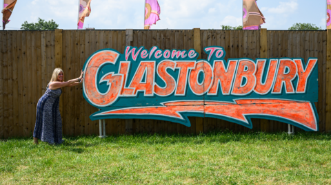 A woman posing next to a sign which says Welcome To Glastonbury
