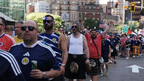Men in blue or red Scotland strip, many in kilts, queue on an American street