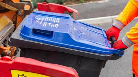 A bin collection worker dressed in hi-vis orange overalls and orange gloves. He is tipping back the handle of a black bin with a blue lid to attach it to the back of a recycling truck.