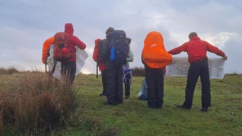 A group of teenagers stood holding a map as they try and navigate around on the moors. They are all wearing red rain jackets and big rucksacks on their backs.