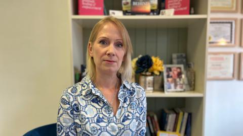 A woman wearing a shirt with a blue and white pattern sat in her home, in front of a bookcase. She has a stern expression.