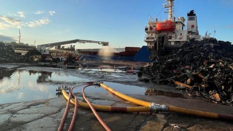 A long-armed aerial platform sprays water over the scene of the fire, with hoses in the foreground and a blue and white ship in the background