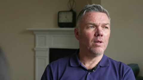 A man with short grey hair wearing a dark blue polo shirt. He is sat in his living room looking off to the right of the image.