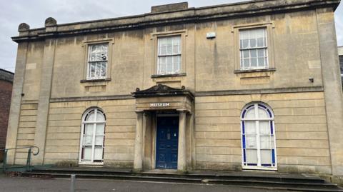 The exterior of Aplsey House. It is a large sand-coloured Georgian building, with two arched windows either side of the door and three rectangular sash windows upstairs. The blue front door has a porch overhang supported by two stone columns. The building appears somewhat run down with graffiti, a smashed window and a dirty façade.