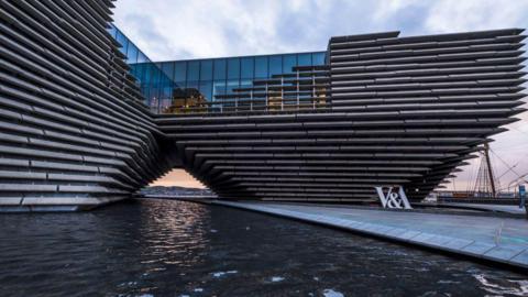 The exterior of the V&A Museum of Design in Dundee. The building features large concrete sections making up its framework. The museum has a large pool of water in front of the building 