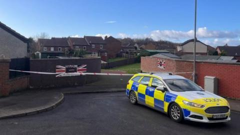 Police attending the scene of the shooting in the Ballyreagh Way area of Newtownards. A police car sits in front of an entry leading to a green area. There is a police cordon in place. The walls of the entry are adorned with union flags.