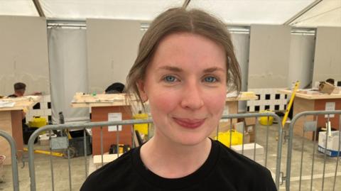 A woman with brown hair, tied up. She is smiling at the camera and is wearing a black top. 