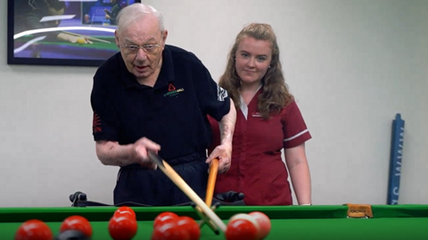 An elderly man in a black polo short take a snooker shot using a rest whilst a young woman in a maroon uniform looks on.