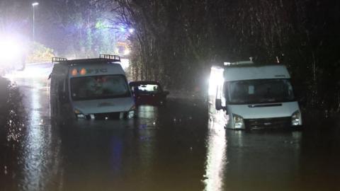 Two white vans and a dark coloured car in between them sit in deep water almost up to the bonnets. Bright lights from emergency vehicles shine in the background in the darkness
