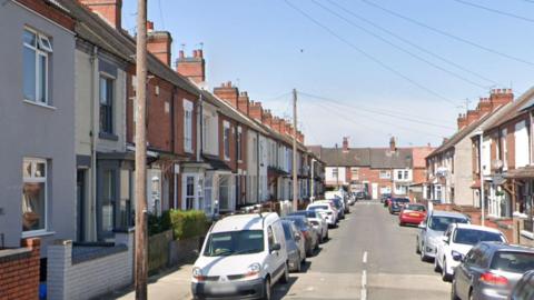 Cheverel Street is a residential road in Nuneaton with terraced houses and parked cars on both sides. There are speed bumps in the road. The houses have gardens with low walls outside. There are lampposts and hedges.