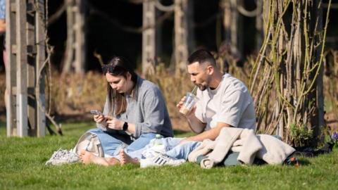 A woman and a man sit in the sunshine on some grass. Both have their trainers off. The man is drinking and iced-coffee and the woman is reading her phone.