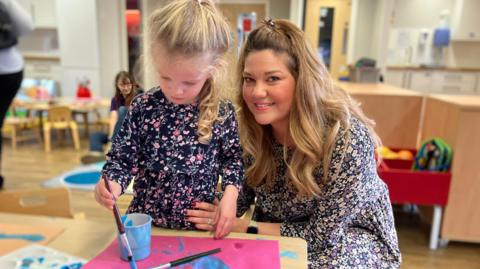 A girl with blonde hair painting while her mum, also with blonde hair, cuddles her.