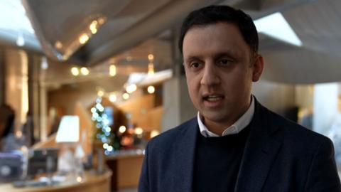 Anas Sarwar stood in the lobby of the Scottish Parliament with blurred lights behind him.