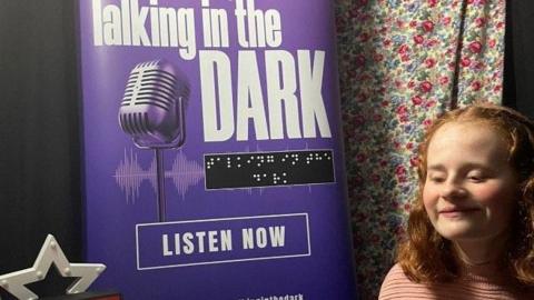 Evie, a teen with ginger hair smiles in front of a purple poster with a microphone on it that says talking in the dark, with braille underneath.