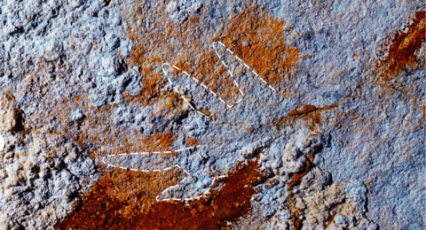 A colour‑enhanced close‑up of a cave wall showing a very faint, claw‑like hand stencil, picked out for clarity with a white dashed outline