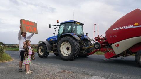 A parent and child hold up a 'thank you' sign as a farmer towing a water tanker passes them