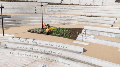 The white stone steps lead to the amphitheatre. There are flower beds dotted around the platforms that break up the steps. The words engraved read: "The town turned round and the Tees; rose up from its bed, performed; its festival, juggled the sun; in its ripples and swirls.; Years of ferrying rust and grief; to the coast, but it stayed true; to its course and its cause; Ask the river the time and it always; says now. Where it jigs and twirls; past these banks the river is ours."