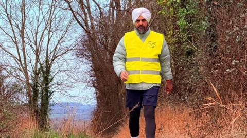 Bhupinder Sandhu walks across the countryside on his second mental health marathon walk. He wears a white turban, a grey weatherproof jacket, blue shorts and leggings, and a hi vis vest.