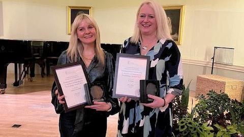 Two woman stand holding photo frames with certificates inside. The pair are also holding awards.