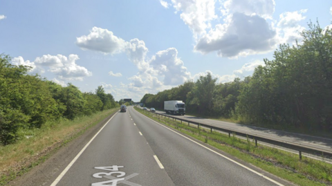 A screenshot from Google street view showing a dual carriageway with a central reservation and barrier in the middle, with trees on either side, on a sunny way.