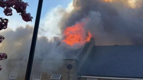 Large bright orange flames can be seen coming out of the top of a rooftop. They are surrounded by thick black smoke