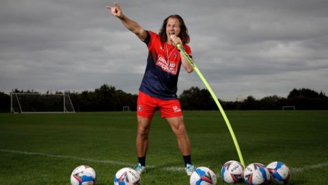 Gareth Ainsworth pretending to sing into a corner flag with footballs scattered around him, while making a 'rock on' hand signal with his right hand, with football pitches in the background 