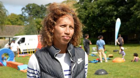 Serena looks at the camera as people set up the sports day behind her. She is wearing a white jumper with blue stripe hoops with a black gillet. Her hair is slightly blowing in the wind.