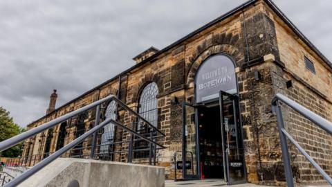 The entrance to Hopetown, a single-storey dark brick building with signage, pictured from the foot of steps leading up to its front entrance There are large, arched windows with black leading.