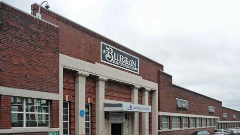 A large redbrick factory with white columns at the front and a tiled sign reading 'Burton the tailor of taste' in black and white above the entrance