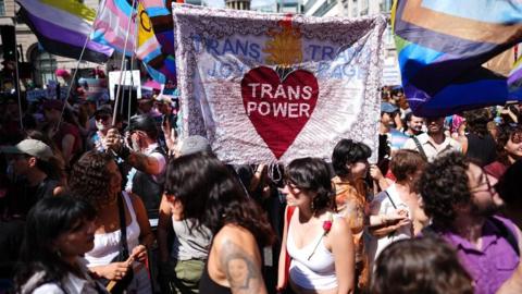 Dozens of people are seen gathered in the background and foreground at the march. A banner is held up by some people in the middle of the photo that reads "Trans joy, trans power, trans rage"