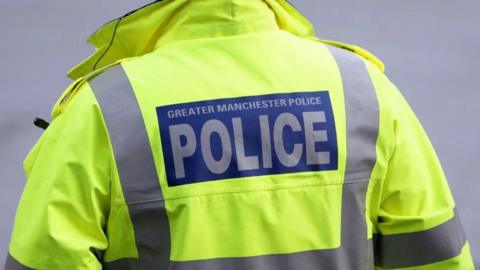 A Greater Manchester Police officer wearing a high-vis yellow jacket facing away in a zoomed in photograph.