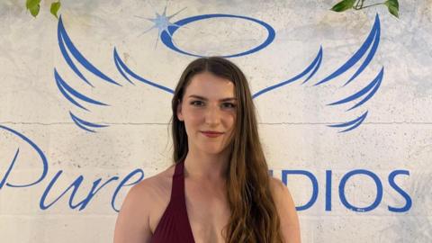 Giselle Pearson at the aerial dance studio. She is standing under a sign that has a halo and wings and there is a studio logo behind her. She is wearing her dance kit. There is foliage decorating the wall.