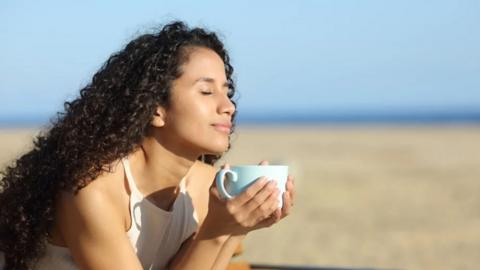 A woman holds a mug while sitting on a sunny beach
