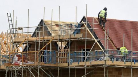 The image shows the upper portion of a building under construction, with scaffolding surrounding the structure. The main focus is on the roof area of the building. Two workers wearing high-visibility safety vests and helmets are visible on the roof.