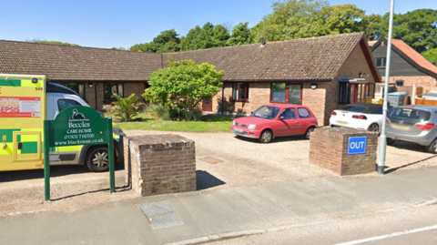 A Google Street View image of the entrance to Beccles Care Home - a small brick building with a dark brown roof. Cars are parked outside and an ambulance can be seen on the left of the image.