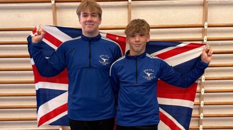 Archie and Samuel in Splitz Gymnastics Club tracksuits standing together holding a Union Jack draped behind them in a gymnasium