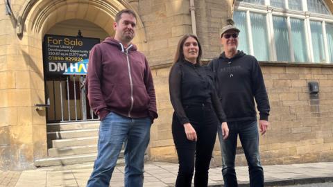 A man with wine-coloured top and blue jeans, a woman dressed in a black top, leggings and boots, and a man in sunglasses and wearing a black hooded top, dark jeans, blue and yellow trainers and a baseball cap stand outside the closed up building library in Jedburgh.