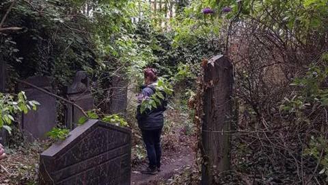 A woman stands in a graveyard looking at stones. There are a lot of trees and shrubs growing around Victorian grave stones.