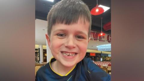 Archie smiles at the camera. He has short dark hair and a big smile on his face, showing a tooth missing from the bottom row. He is wearing a navy football shirt with yellow trim and appears to be in a restaurant