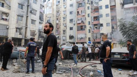 People inspect the site of an Israeli strike in the southern suburbs of Beirut a car has been overturned and there is visible damage to flats