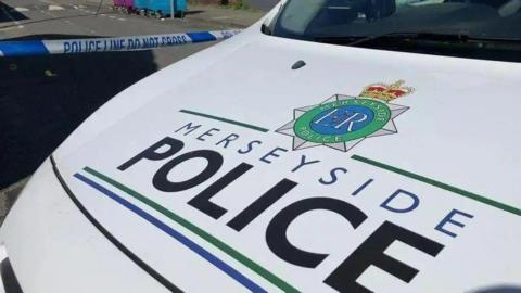 A Merseyside Police van with the force's name and logo spread over the white bonnet. Police tape can be seen pulled over a road in the background. 