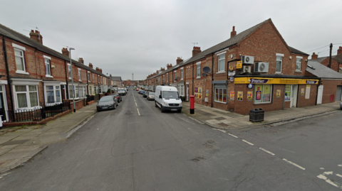 The street has red bricked terraced houses on either side. On the left, there is a yellow sign for the 'Premier' convenience store